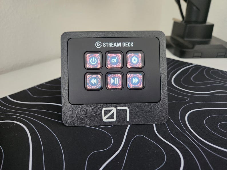 May include: Black Stream Deck with six illuminated buttons featuring various icons. The device has the "Stream Deck" logo and a white graphic. It sits on a black and white patterned surface.