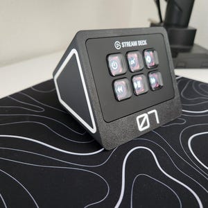 May include: Black Stream Deck with six illuminated buttons and the Stream Deck logo. The device is on a black and white topographic patterned mousepad. The number "07" is printed on the front.