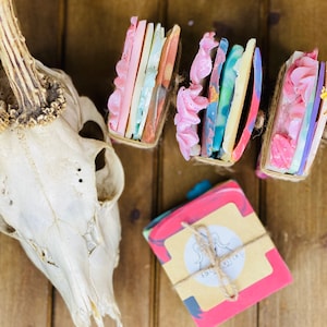 May include: Four bars of handmade soap wrapped in twine, with a fifth bar of soap wrapped in twine and a label that says "Soap Sisters". The soaps are various colors, including pink, blue, green, and yellow.