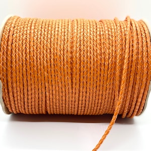 May include: A spool of orange braided leather cord. The cord is wound around a cardboard spool and a length of cord is hanging off the spool.