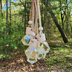 May include: A collection of hanging car air fresheners. Each freshener features a clear, geometric-shaped glass bottle with a wooden cap and a beige rope for hanging. The bottles are filled with a clear liquid. The background is a blurred forest scene.