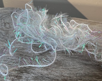 May include: White, iridescent yarn with a fuzzy texture. The yarn has a shiny, reflective quality with strands of green, purple, and blue. The yarn is loosely coiled and appears soft, suitable for crafting projects.