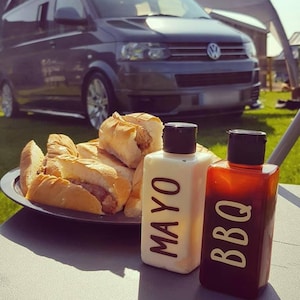 May include: A close-up of two condiment bottles labeled "MAYO" and "BBQ" on a table with a plate of hot dogs in the background.