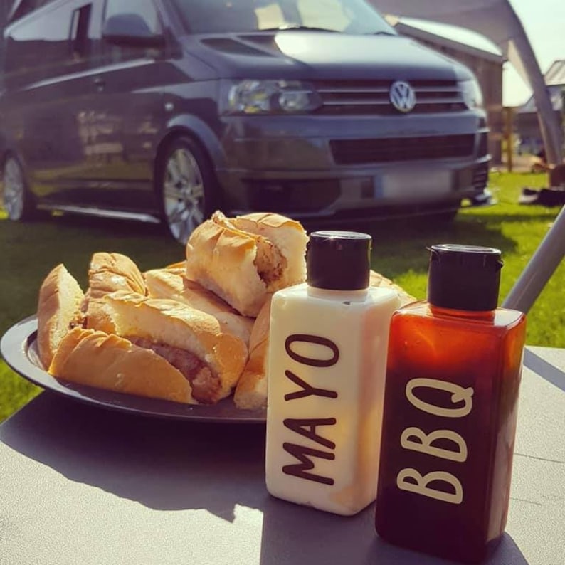 May include: A close-up of two condiment bottles labeled "MAYO" and "BBQ" on a table with a plate of hot dogs in the background.