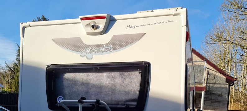 K&ouml;nnte beinhalten: Ein wei&szlig;er Wohnwagen mit einem grauen und silbernen "Signature"-Logo und dem Text "Making memories one road trip at a time..." auf der R&uuml;ckseite. Der Wohnwagen hat ein Heckfenster mit einem grauen Netzgitter und eine R&uuml;ckfahrkamera.