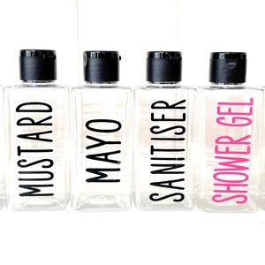 May include: Eight clear plastic bottles with black pump tops and labels. The labels have black and pink text. The labels read "MAYO", "WASHING UP", "MUSTARD", "MAYO", "SANITISER", "SHOWER GEL", "SHAMPOO", and "CONDITIONER".