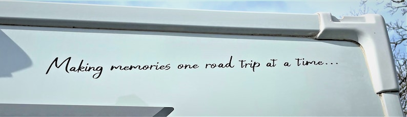 K&ouml;nnte beinhalten: Ein wei&szlig;er Wohnwagen mit dem Text "Making memories one road trip at a time..." in schwarzer Kursivschrift an der Seite.