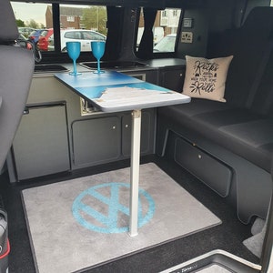 K&ouml;nnte beinhalten: Ein graues und schwarzes Wohnmobil-Interieur mit einem Klapptisch, zwei blauen Gl&auml;sern und einem grauen Teppich mit einem blauen Volkswagen-Logo. Der Text "The Rocks where your home Rolls" befindet sich auf einem wei&szlig;en Kissen auf der Sitzbank.