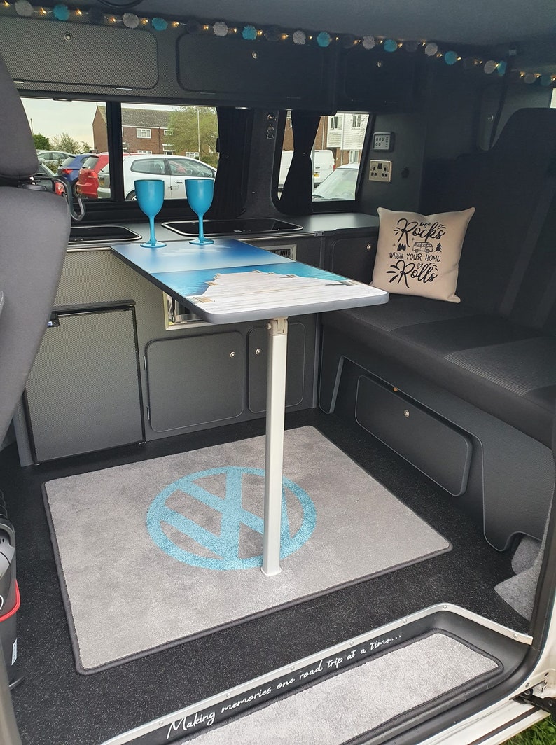 K&ouml;nnte beinhalten: Ein graues und schwarzes Wohnmobil-Interieur mit einem Klapptisch, zwei blauen Gl&auml;sern und einem grauen Teppich mit einem blauen Volkswagen-Logo. Der Text "The Rocks where your home Rolls" befindet sich auf einem wei&szlig;en Kissen auf der Sitzbank.