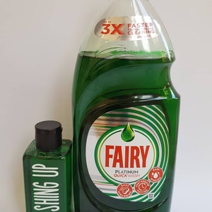 May include: Two bottles of Fairy Platinum Quick Wash dish soap. The larger bottle is green and has a red cap. The smaller bottle is green and has a black cap. The larger bottle has a label that says "Up to 3X Faster Tough Grease Cleaning". The smaller bottle has a label that says "Washing Up".