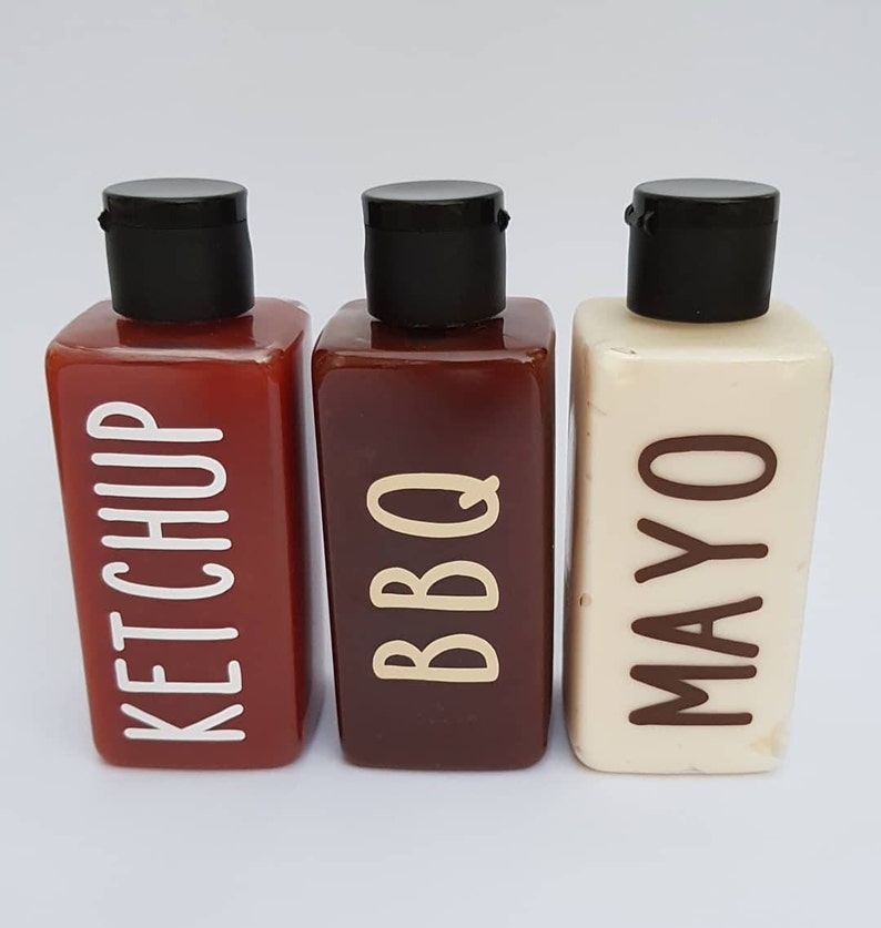 May include: Three clear plastic bottles with black lids and labels. The bottles are filled with red, brown, and white liquids. The labels read "Ketchup", "BBQ", and "Mayo".