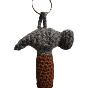 May include: A handmade crochet hammer keychain. The hammer head is gray, the handle is brown, and the connecting piece is gray. The keychain has a silver metal ring for attaching to keys or bags. The hammer is a novelty item.