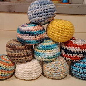 May include: A collection of ten round, crocheted hacky sacks stacked in a pyramid formation. The items feature various yarn colors including solid yellow, white, brown, and multi-colored variegated patterns in shades of blue, red, and orange.