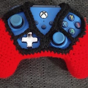 May include: A red and black crocheted cover for a blue Xbox controller. The cover has a black crocheted design and red crocheted sides.