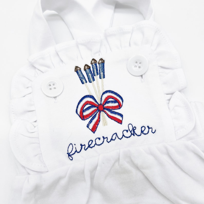 May include: White sleeveless romper with a red, white, and blue bow and firework design. The romper has a ruffle trim and says "firecracker" in blue embroidery.