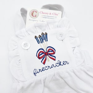 May include: White sleeveless romper with a red, white, and blue embroidered design of fireworks and a bow. The text "firecracker" is embroidered below the design.