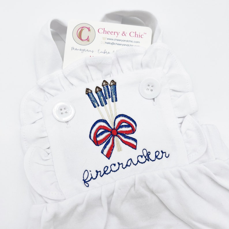 May include: White sleeveless romper with a red, white, and blue embroidered design of fireworks and a bow. The text "firecracker" is embroidered below the design.