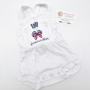 May include: White baby onesie with a red, white, and blue embroidered design of fireworks and a bow with the word "firecracker" underneath.