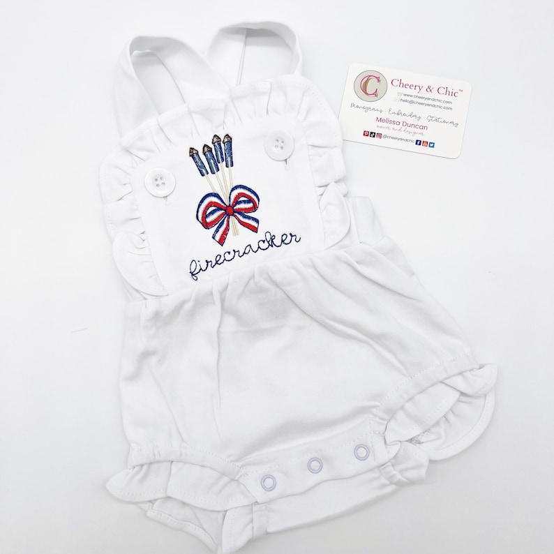 May include: White baby onesie with a red, white, and blue embroidered design of fireworks and a bow with the word "firecracker" underneath.