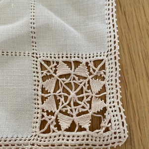 May include: A white linen square tablecloth with a delicate lace border. The lace is a cream color and features a geometric pattern.