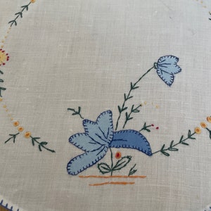 Puede incluir: Un mantel redondo de lino blanco con flores bordadas azules y amarillas. El mantel tiene un borde festoneado azul.