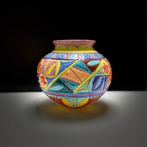 Puede incluir: Un jarrón de cerámica colorido con un diseño geométrico en tonos de azul, verde, amarillo, naranja, rojo y morado. El jarrón tiene una forma redondeada y un acabado liso y brillante.