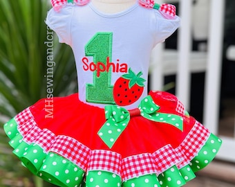May include: A white t-shirt with a green number 1 and the name "Sophia" in red, paired with a red tutu skirt. The skirt features green polka dot and red gingham accents, with matching bows on the shoulders and skirt.