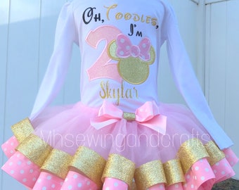 May include: A white long-sleeved shirt and pink tutu set for a toddler. The shirt has the text "Oh Coodles, I'm 2 Skylar" with a Minnie Mouse design. The tutu is pink with gold and polka dot accents.
