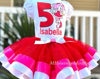 May include: A white t-shirt with a large red number 5 and the name "Isabella" in white, paired with a red, pink, and white tutu skirt. The shirt features a cartoon character. The outfit is designed for a child's birthday.