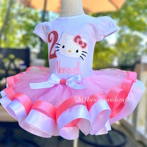 May include: A pink and white tutu with a white shirt featuring a Hello Kitty design and the number 2. The shirt also has the name "Nevaeh" embroidered on it.