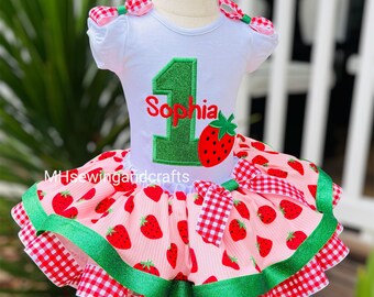 May include: A white short-sleeved dress with a strawberry-themed tutu skirt. The dress features a large green number "1" and the name "Sophia" in red. The skirt has a pink background with strawberry patterns, red gingham trim, and green ribbon accents.