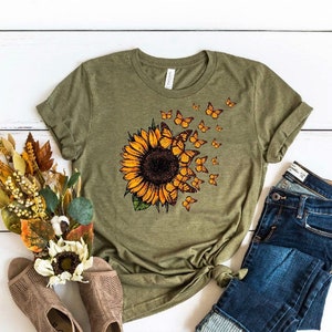 K&ouml;nnte beinhalten: Ein gr&uuml;nes T-Shirt mit einem Sonnenblumen- und Schmetterlingsmotiv. Der Text "H. Olive" ist unten auf dem Shirt aufgedruckt.