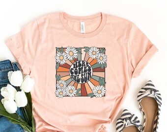 May include: A peach colored t-shirt with a retro graphic design featuring a sun with rays and the text "Here Comes The Sun" surrounded by daisies. The t-shirt is on a white background with a pair of blue jeans and white flowers.