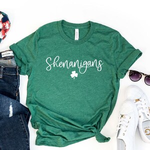 May include: A green t-shirt with the word "Shenanigans" and a shamrock printed in white. The shirt is on a white background with a pair of jeans, a headband, sunglasses, and white sneakers.