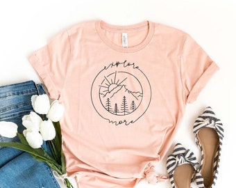 May include: A heather peach colored t-shirt with a black graphic of a mountain, sun, and trees in a circle with the text "explore more".