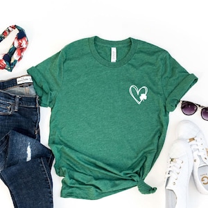 Op de afbeelding: Een groen T-shirt met een wit hart en klavertje vier design. Het shirt wordt gecombineerd met een blauwe jeans, een bloemen hoofdband, witte sneakers en een zonnebril.