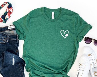 Puede incluir: Una camiseta verde con un corazón blanco y un diseño de trébol. La camiseta se combina con jeans azules, una diadema floral, zapatillas blancas y gafas de sol.