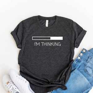 May include: A dark grey t-shirt with a white graphic that reads "I'M THINKING" and a progress bar. The shirt is laid flat on a white surface with a pair of white sneakers and blue jeans.
