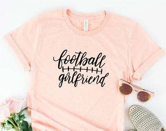 May include: A pink t-shirt with the text "football girlfriend" in black script with a football field line graphic underneath. The shirt is laid flat on a white surface with a pair of sunglasses and a pair of white shoes.