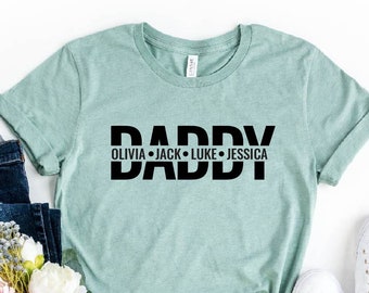 May include: A light blue t-shirt with the word "Daddy" in black letters and the names "Olivia, Jack, Luke, Jessica" underneath. The shirt is on a white background with a pair of jeans, a bouquet of flowers, and a pair of white shoes.