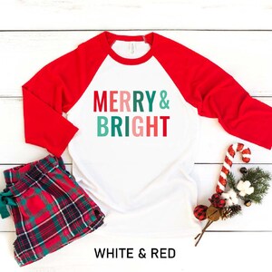 Puede incluir: Camiseta de b&eacute;isbol blanca y roja con el texto "Merry & Bright" en verde, rosa y azul. La camiseta est&aacute; sobre un fondo de madera blanca con pantalones de pijama a cuadros rojos y verdes, un bast&oacute;n de caramelo y una rama de vegetaci&oacute;n.
