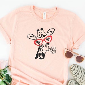 May include: A heather peach coloured t-shirt featuring a black and white giraffe wearing red glasses and holding a flower. The text "HEATHER PEACH" is at the bottom of the image.