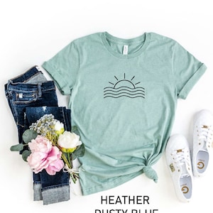 May include: A heather dusty blue t-shirt featuring a simple black graphic of a sun setting over wavy lines. The shirt is styled with blue jeans, a bouquet of pink flowers, and white trainers.