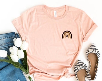 May include: A peach colored t-shirt with a rainbow graphic on the left chest. The rainbow is made of brown, black, and beige stripes with a small heart in the center. The t-shirt is laid flat on a white background with a pair of blue jeans, a bouquet of white tulips, and a pair of black and white striped flats.