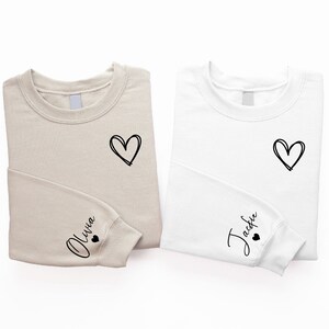 Peut inclure: Deux sweats beige et blanc avec un c&oelig;ur et un nom sur chacun. Le sweat beige a "Olivia" sur la manche et le sweat blanc a "Jackie" sur la manche.