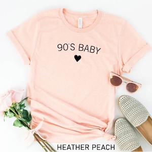 May include: A light pink t-shirt with the text "90's Baby" and a black heart. The shirt is made of a soft, comfortable material and has a relaxed fit.