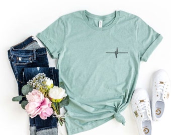 Könnte beinhalten: Ein T-Shirt in der Farbe „Heather Dusty Blue“ mit einer einfachen schwarzen Herzschlaggrafik auf der Vorderseite. Das Shirt wird mit einer blauen Jeans und weißen Turnschuhen kombiniert.