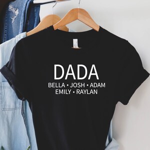 May include: Black t-shirt with white text that reads "DADA" and the names "BELLA, JOSH, ADAM, EMILY, RAYLAN" underneath. The shirt is hanging on a hanger.