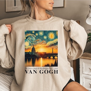 May include: A beige sweatshirt with a print of a painting of a cityscape at night with a starry sky. The painting is titled "The Starry Night in Washington, D.C., 1889" and signed "VAN GOGH".