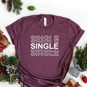 May include: Heather maroon t-shirt with the word "SINGLE" stacked in white block letters. The shirt is surrounded by holiday decorations, including wrapped gifts, pine branches, and ornaments.